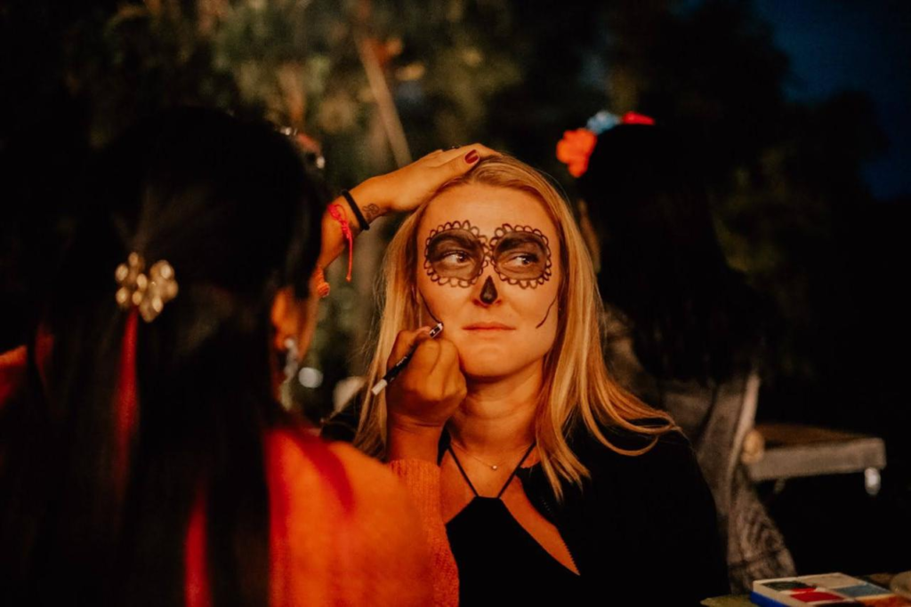 A woman sits patiently as an artist applies intricate Day of the Dead face paint to her. The makeup features dark eye circles and a nose design resembling a sugar skull pattern. The scene is set outdoors at night, with soft lighting and a relaxed atmosphere. The artist, partially visible from behind, carefully works on the details, holding the woman's face for precision.