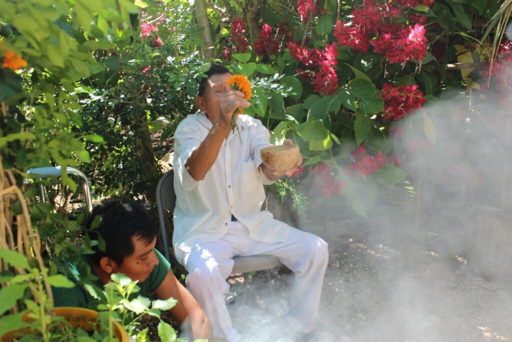 An elderly man dressed in white performs a traditional ceremony outdoors, holding a small bowl in one hand and a marigold flower in the other. He sits in a lush garden filled with green plants and vibrant red flowers, with smoke rising from the ceremony, creating a mystical atmosphere. A younger man is kneeling nearby, focusing on the ritual. The setting suggests a spiritual or cultural practice, possibly related to Hanal Pixán or Day of the Dead traditions.